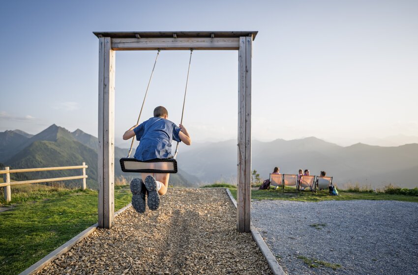 Child swings on wooden swing on a mountain overlooking the Alps, with people sitting in the background | © Dorfgasteiner Bergbahnen AG