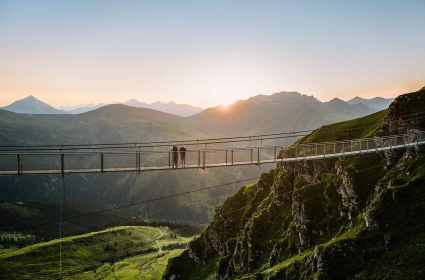 Two people on Stubnerkogel suspension bridge at sunset with view of green mountains and Gastein Valley | © Gasteiner Bergbahnen AG