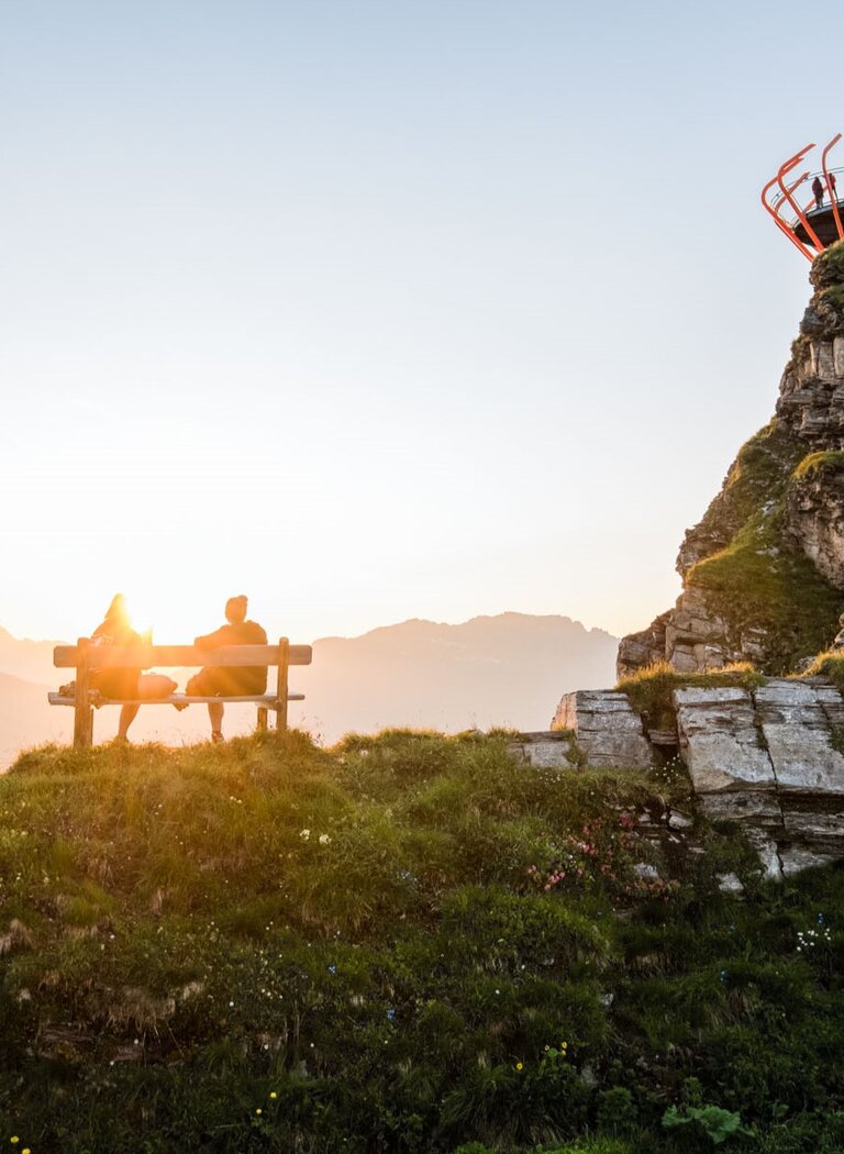 Couple on bench at sunset facing mountains, with Glocknerblick platform on grassy rock to the right | © Gasteiner Bergbahnen AG
