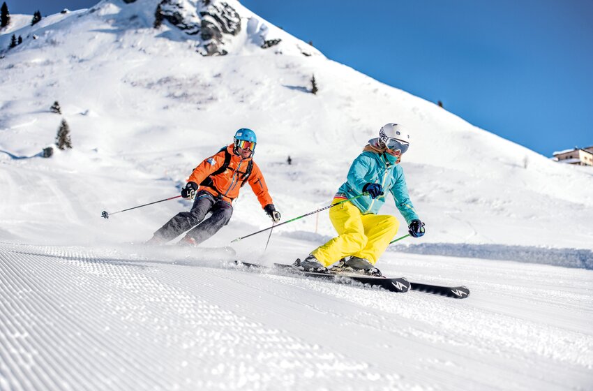 Two skiers carve down a wide, freshly groomed slope in sunshine and powder snow. | © Gasteiner Bergbahnen AG