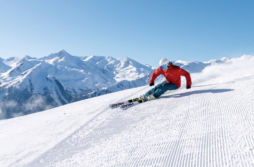 Skier in red jacket carving on freshly groomed slope with snowy peaks and blue sky in the background. | © Gasteiner Bergbahnen AG