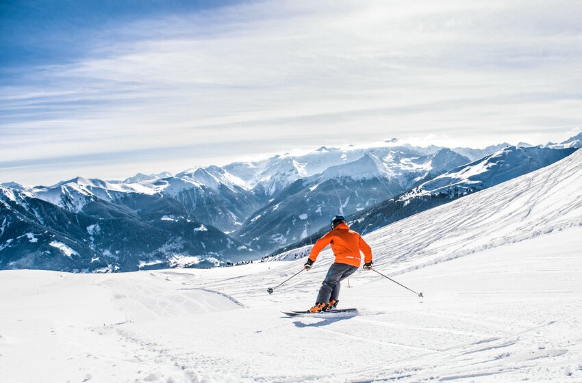 Skier in orange jacket descends a wide slope with views of snow-covered valleys and alpine peaks.  | © Gasteiner Bergbahnen AG