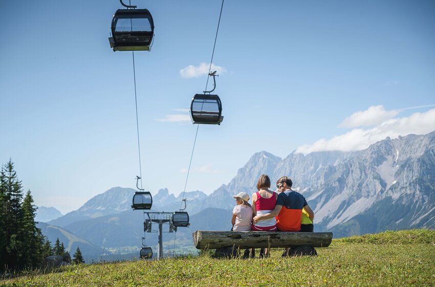 Family sitting on a bench overlooking gondolas and the impressive Dachstein mountain range. | © Ski amadé GmbH