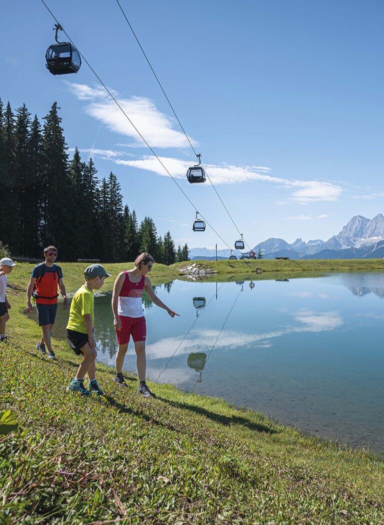 Family walking by a clear alpine lake, gondolas above and mountain panorama in background under blue sky. | © Ski amadé GmbH