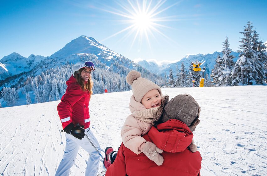 Woman with skis and child in winter clothes on a ski slope, with bright sun and snowy mountains in the background. | © Ski amadé GmbH