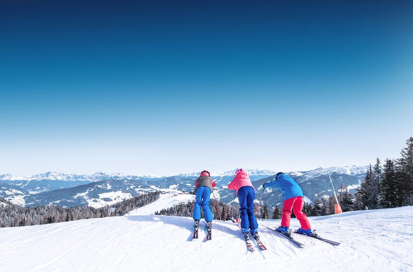 Three children at the top of a ski slope, ready to go downhill with snowy mountains in the background. | © Ski amadé GmbH