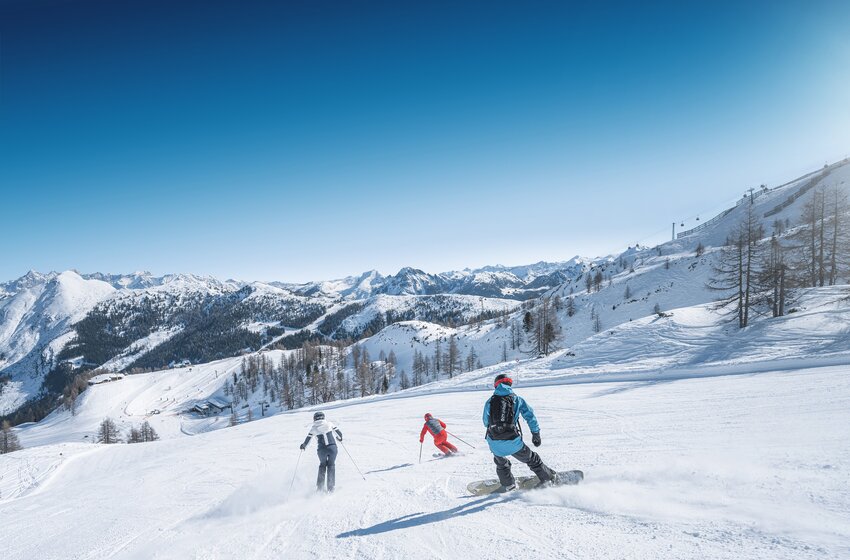 Three winter sports fans – two skiers and one snowboarder – ride sunlit wide slopes with scenic mountain views in Ski amadé. | © Ski amadé GmbH