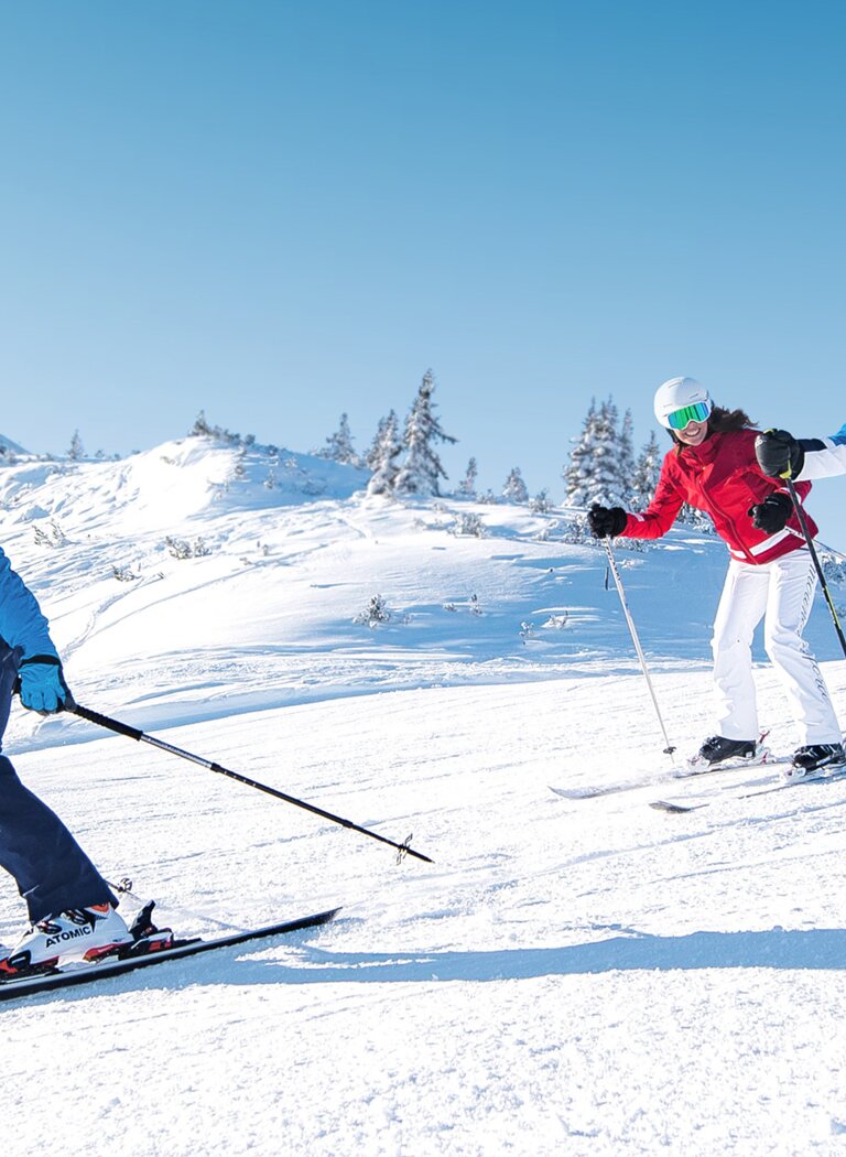 Three skiers skiing side by side on a sunny, snowy slope with chairlifts in the background in Ski amadé. | © Ski amadé GmbH