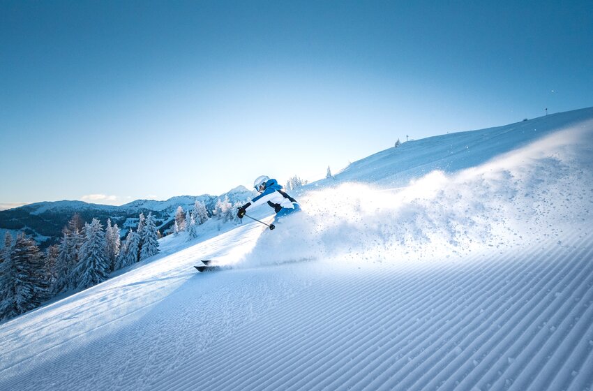 A skier carves powerful turns through fresh powder snow on a sunny, freshly groomed slope with forest backdrop in Ski amadé. | © Ski amadé GmbH
