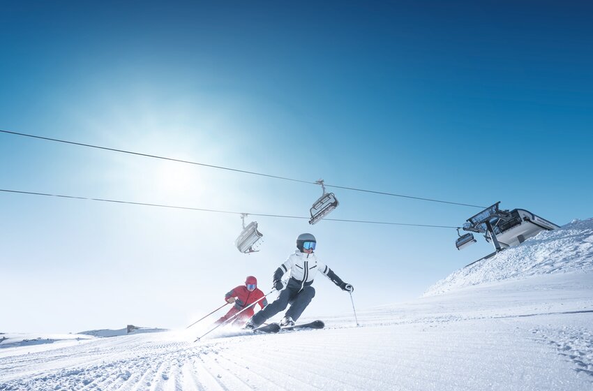 Two skiers carve on a freshly groomed slope beneath a chairlift under a bright blue sky and glowing morning sun. | © Ski amadé GmbH