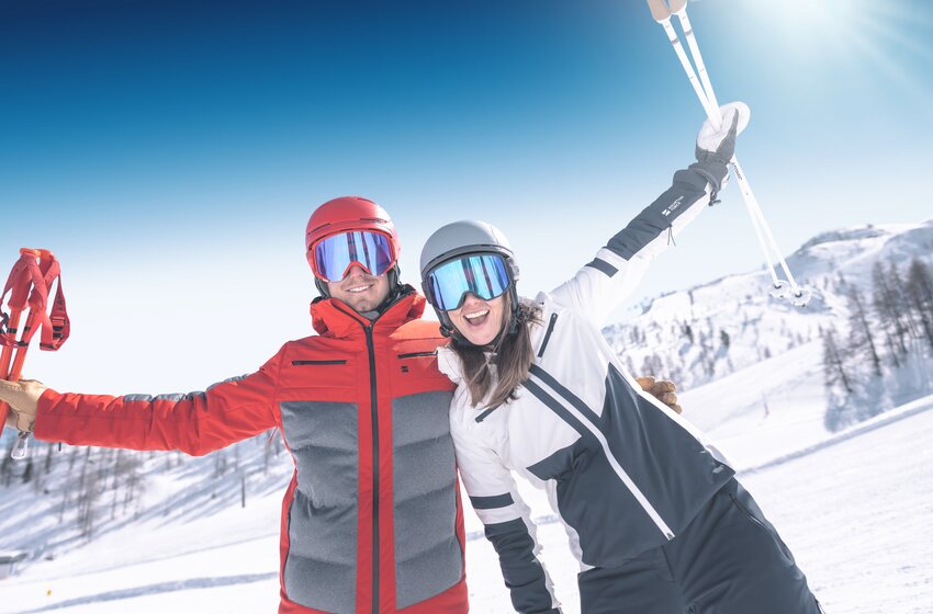 Two smiling skiers stand side by side, arms raised, wearing helmets and goggles against a sunny snowy mountain backdrop. | © Ski amadé GmbH