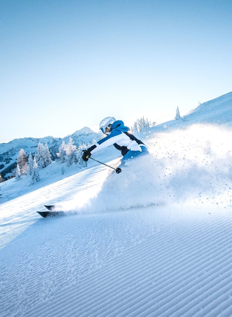 Ein Skifahrer zieht bei Sonnenlicht Schwünge durch frischen Schnee und wirbelt Pulverschnee auf einer Piste in Ski amadé auf. | © Ski amadé GmbH