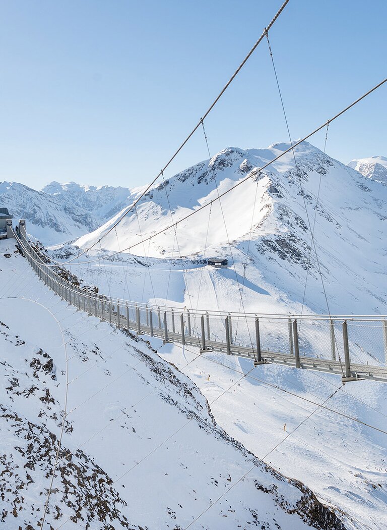 Two people cross a suspension bridge above snow-covered mountains near a mountain station under clear sky | © Gasteiner Bergbahnen AG