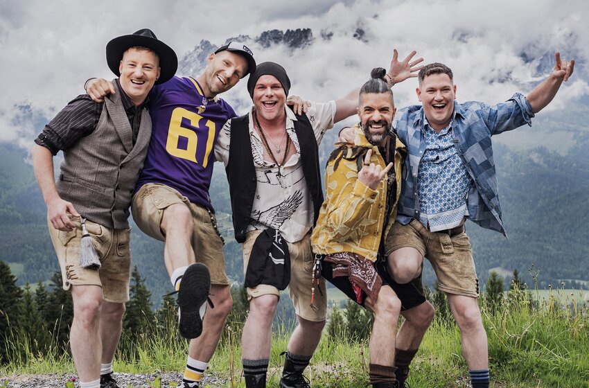 Fünf Männer in Lederhosen posieren Arm in Arm auf einer Wiese vor bewölkten, felsigen Bergen. | © Angelo Lair