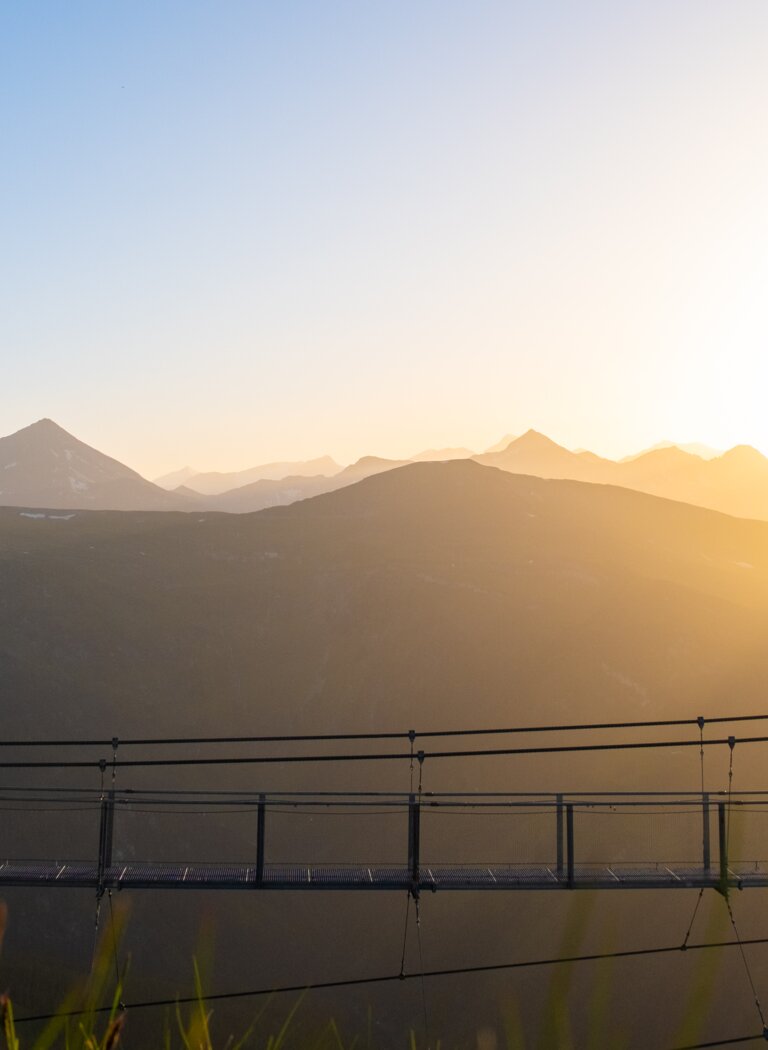 Menschen überqueren Hängebrücke im Sonnenuntergang, im Hintergrund Bergpanorama mit schneebedeckten Gipfeln | © Gasteiner Bergbahnen AG