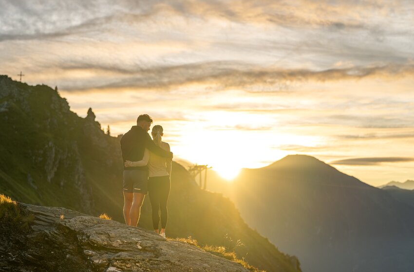 Couple stands closely embraced on a rock at the mountain summit, mountains and glowing sky in background | © Gasteiner Bergbahnen AG