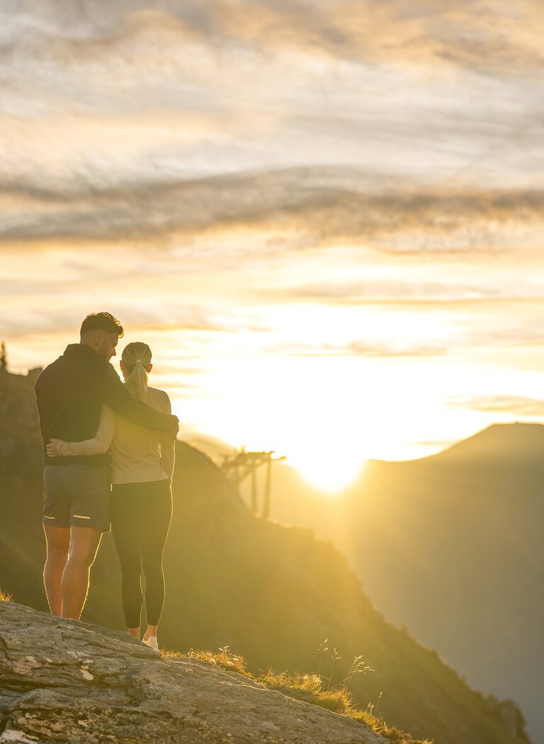 Paar steht eng umschlungen auf einem Felsen am Berggipfel, im Hintergrund Berge und leuchtender Himmel | © Gasteiner Bergbahnen AG