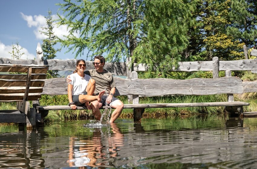 Couple sits on wooden bench by pond dipping feet in water, splashes visible, surrounded by trees and fence | © Gasteiner Bergbahnen AG