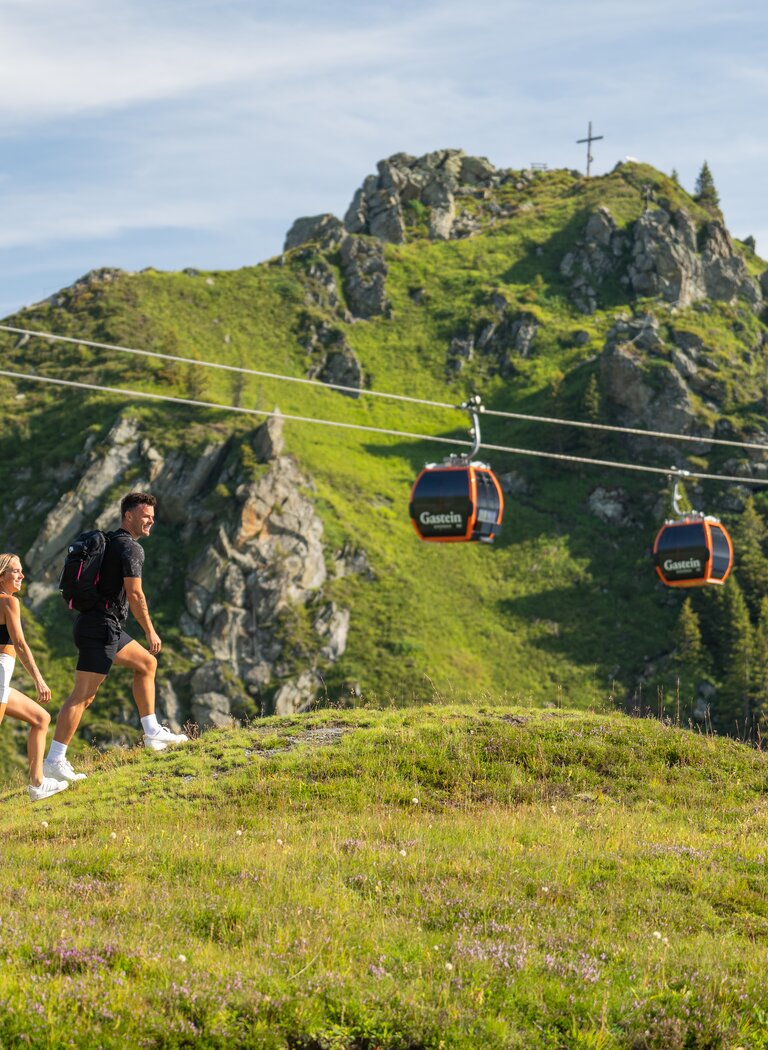 Paar wandert über grüne Almwiese mit Rucksack, im Hintergrund Gondeln und felsiger Gipfel mit Kreuz | © Gasteiner Bergbahnen AG