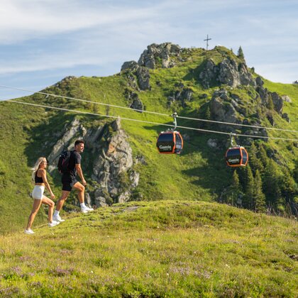 Couple hiking across green alpine meadow with backpack, gondolas behind and rocky summit with cross | © Gasteiner Bergbahnen AG