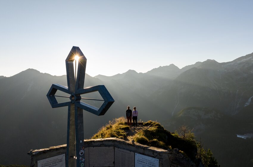 Two hikers stand by a summit cross on a ridge, sun shining through metal cross, alpine mountains in background | © Gasteiner Bergbahnen AG