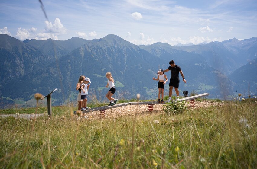 Family with children balancing on wooden play equipment on alpine meadow, mountain panorama and valley in background | © Gasteiner Bergbahnen AG