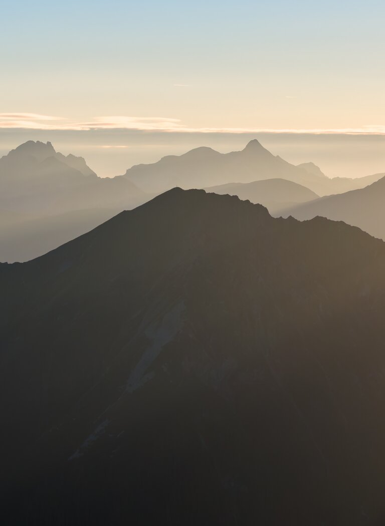 Multiple mountain ranges at sunrise in backlight, soft layers of mist lie between peaks under a clear sky | © Gasteiner Bergbahnen AG