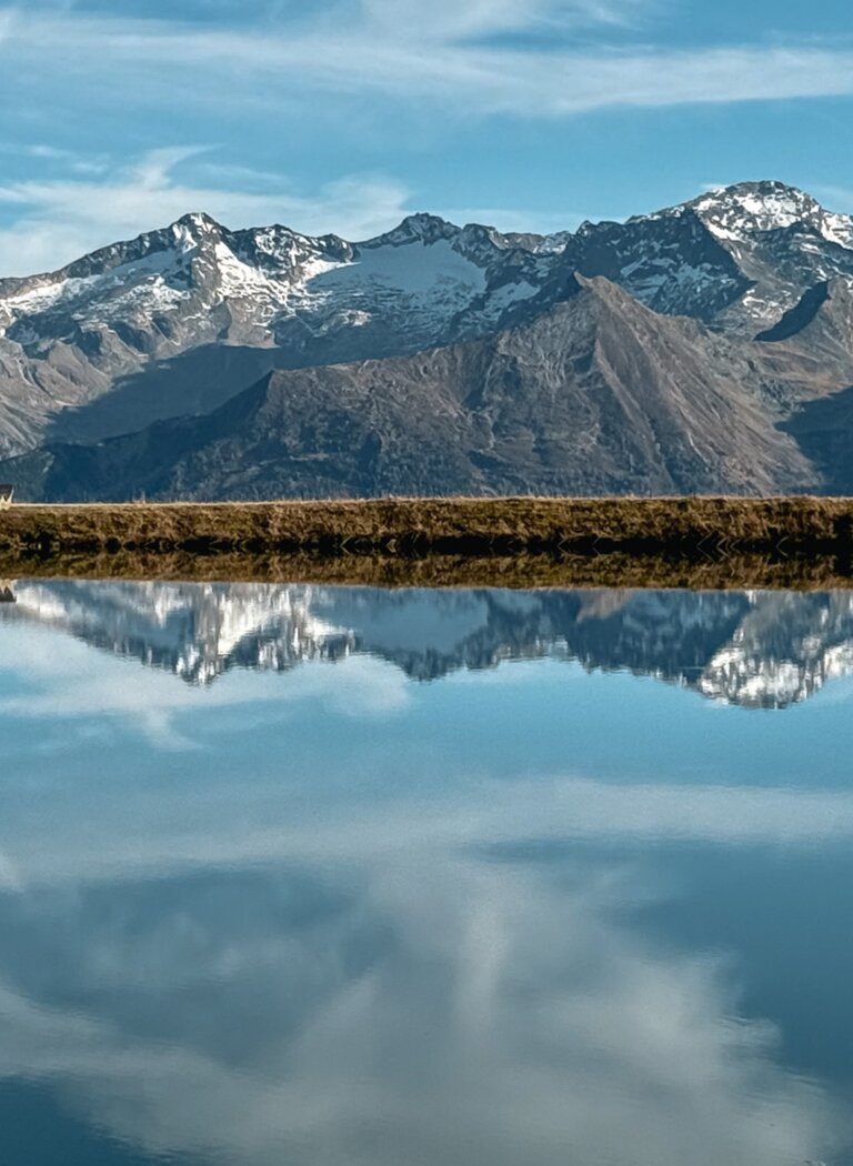 Calm mountain lake reflects snow-capped peaks and blue sky with clouds, a narrow strip of land runs along the shore | © Gasteiner Bergbahnen AG