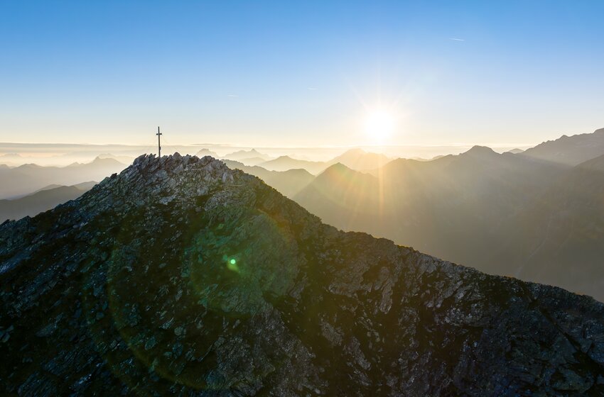 Felsiger Berggrat mit Gipfelkreuz im Morgenlicht, Sonne überlagertem Gebirge und Dunst in den Tälern | © Gasteiner Bergbahnen AG