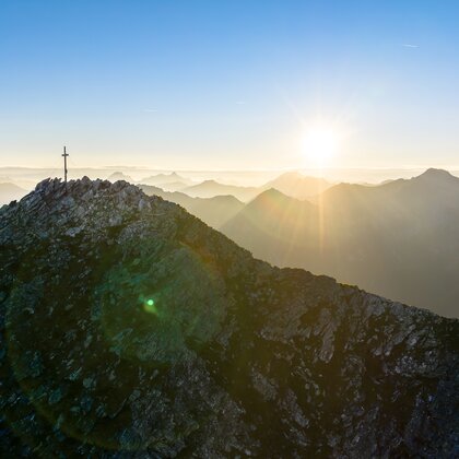 Rocky mountain ridge with summit cross at sunrise, sun over layered ranges and haze in the valleys | © Gasteiner Bergbahnen AG