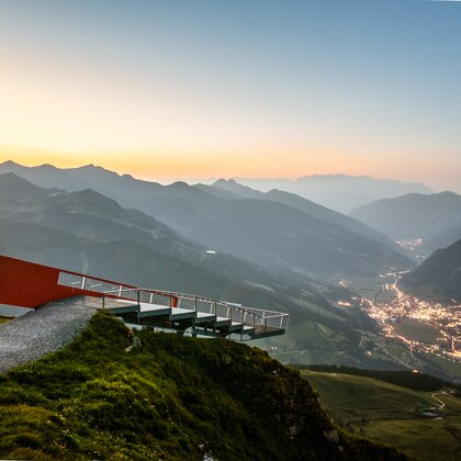 Moderne Aussichtsplattform auf Bergkante über beleuchtetem Tal mit Orten und Serpentinenstraße im Abendlicht | © Gasteiner Bergbahnen AG