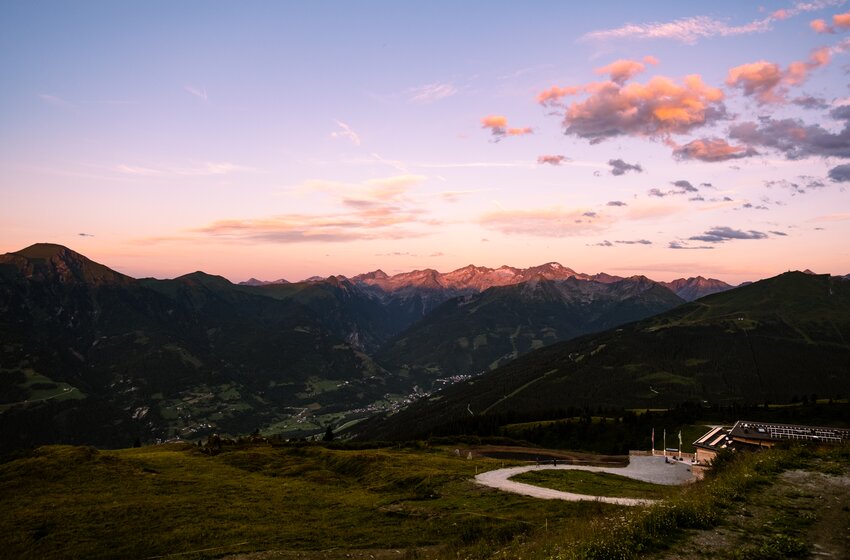 Berglandschaft bei Sonnenuntergang mit grünem Tal, Serpentinenweg im Vordergrund und Gebirgskette am Horizont | © Gasteiner Bergbahnen AG