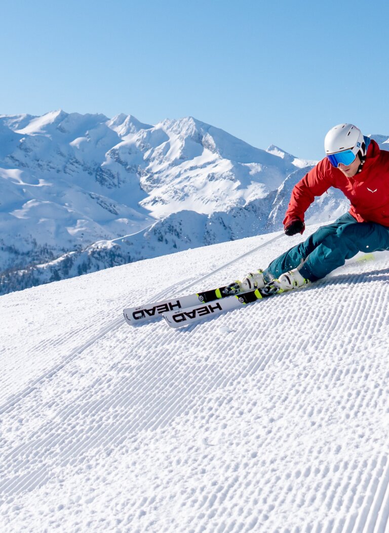 Ein Skifahrer zieht einen Carving-Schwung auf perfekt präparierter Piste vor verschneiten Alpengipfeln. | © Christoph Oberschneider
