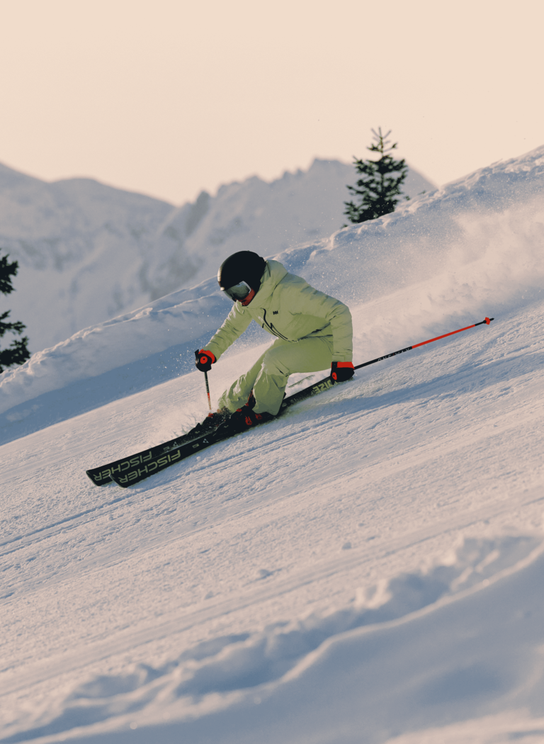 Skier in light clothing carves a turn on a snowy piste, with fir trees and snow-covered mountains in the background. | © Gasteiner Bergbahnen AG 